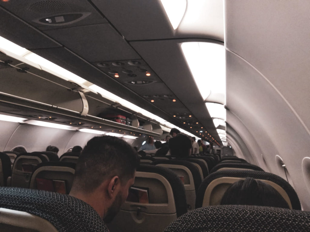 A imagem mostra o interior do avião que peguei para ir até Montevidéu. Aparecem os topos dos assentos da aeronave, os detalhes do topo do interior do avião e alguns dos passageiros