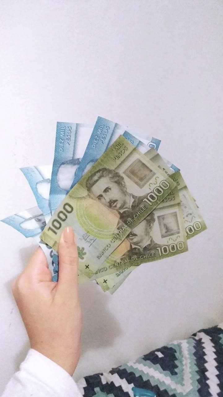 Mão segurano nove notas de pesos chilenos: cinco notas são azuis, de dez mil pesos, e quatro são verdes, de mil pesos cada. O fundo é branco
