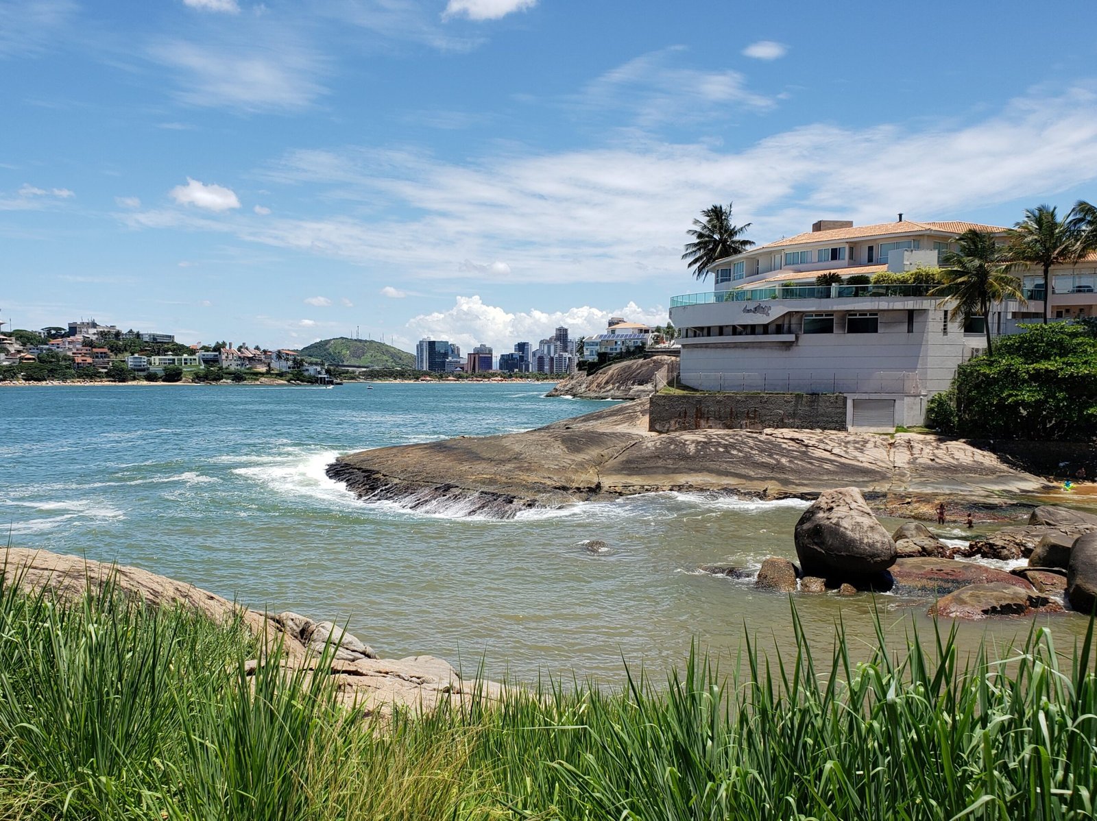 Praia da Castanheira em Vitória, Espírito Santo, com a cidade ao fundo, uma grande casa no canto direito e o mar em tons de azul e verde atrás da vegetação