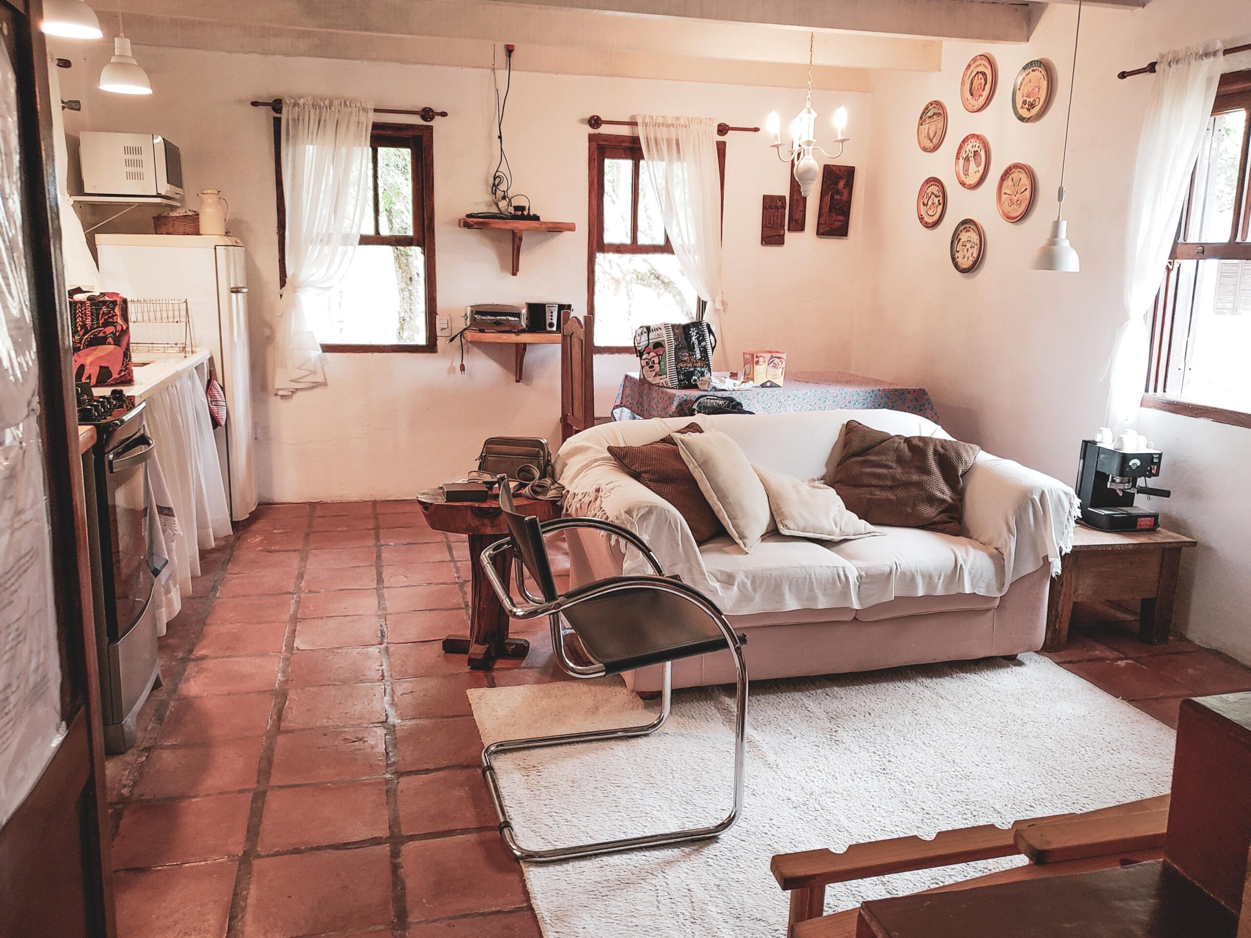Vista da sala, cozinha e sala de jantar do Airbnb em Monte Verde. Em primeiro plano, o sofá está virado para a frente, no canto esquerdo, com uma cadeira a sua frente e um tapete claro no meio. À direita fica a cozinha, enquanto a mesa de jantar, com pratos pendurados como quadros na parede, está diretamente atrás do sofá