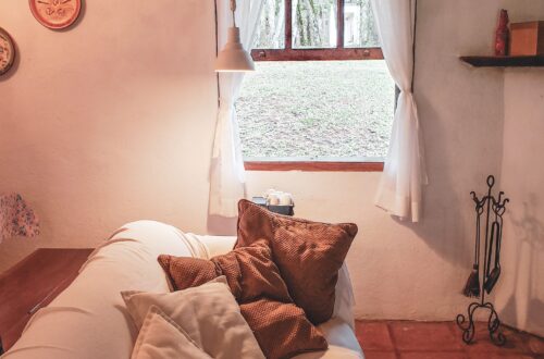 Sala do Airbnb em Monte Verde, com o sofá claro em primeiro plano e a janela de madeira ao fundo