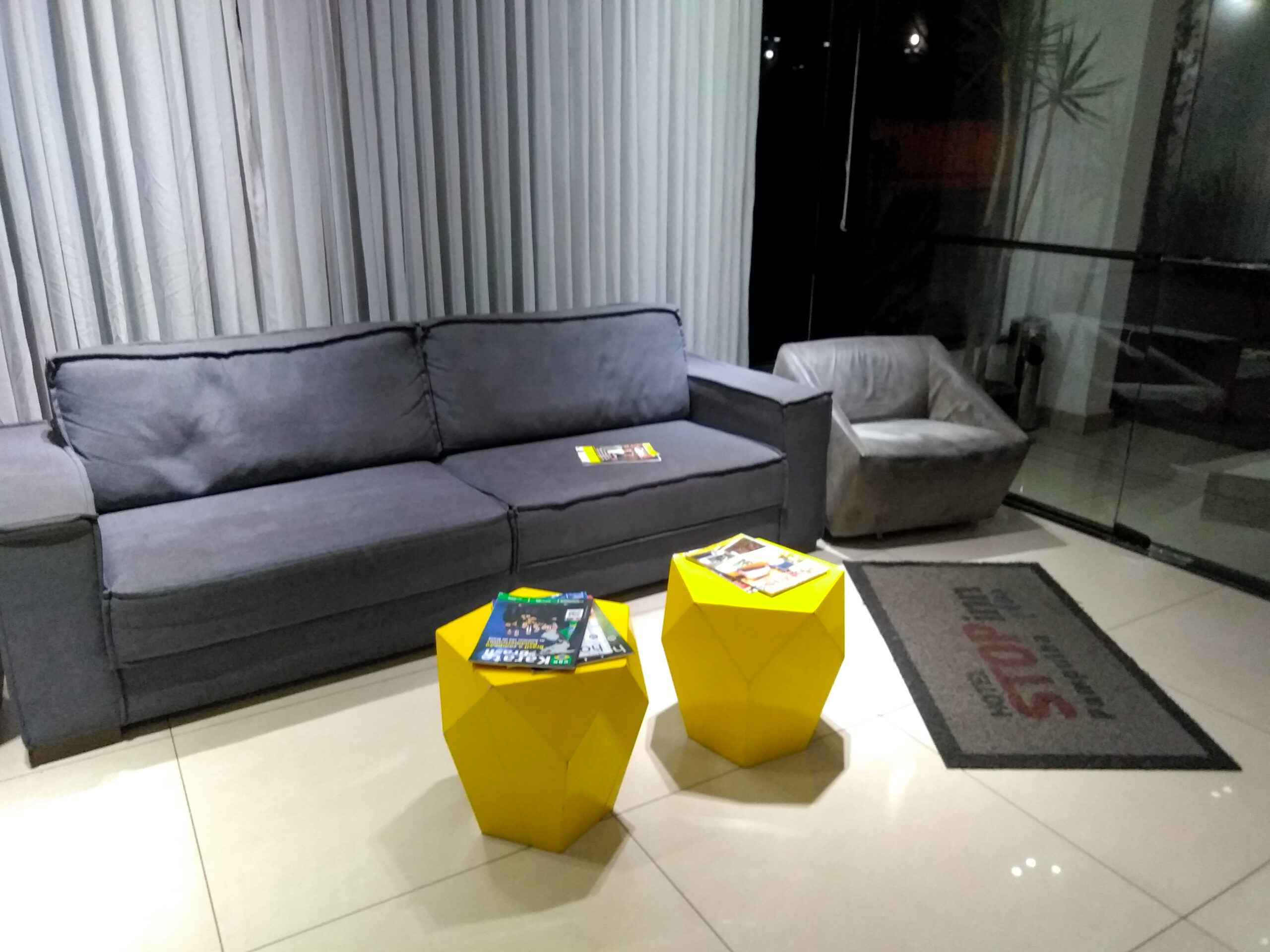 Recepção do Stop Inn Plus Pampulha, com sofá cinza, poltrona cinza clara, mesinhas de centro amarelas e cortina branca