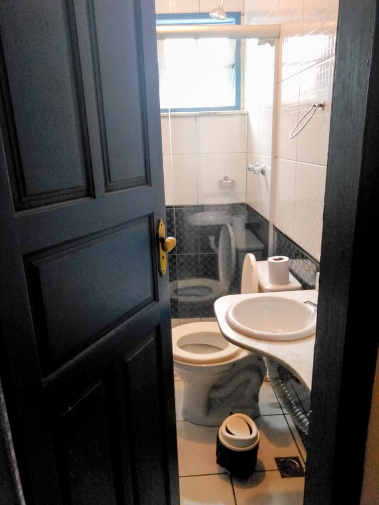 Banheiro da pousada, com porta azul, box, vaso sanitário e pequena pia