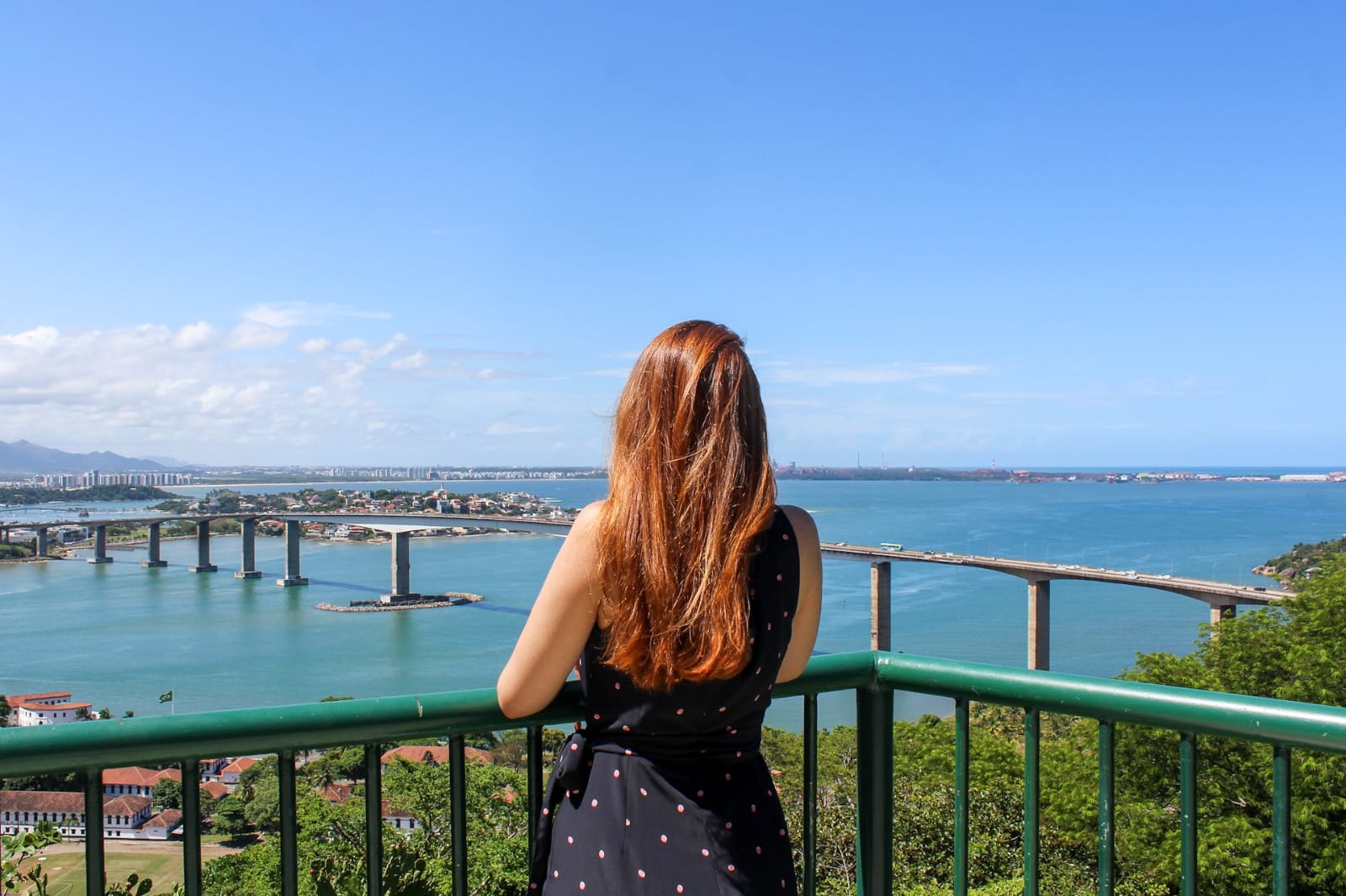 Mulher ruiva de costas, no mirante do Convento da Penha, com vista para a Baía e a Terceira Ponte, com céu azul