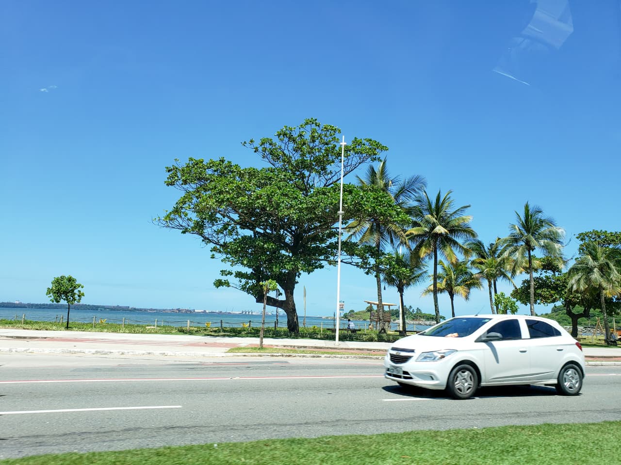 Carro branco na Orla de Camburi, Mata da Praia, em Vitória. Com árvores no canto direito, céu azul e um pouco do mar no canto esquerdo
