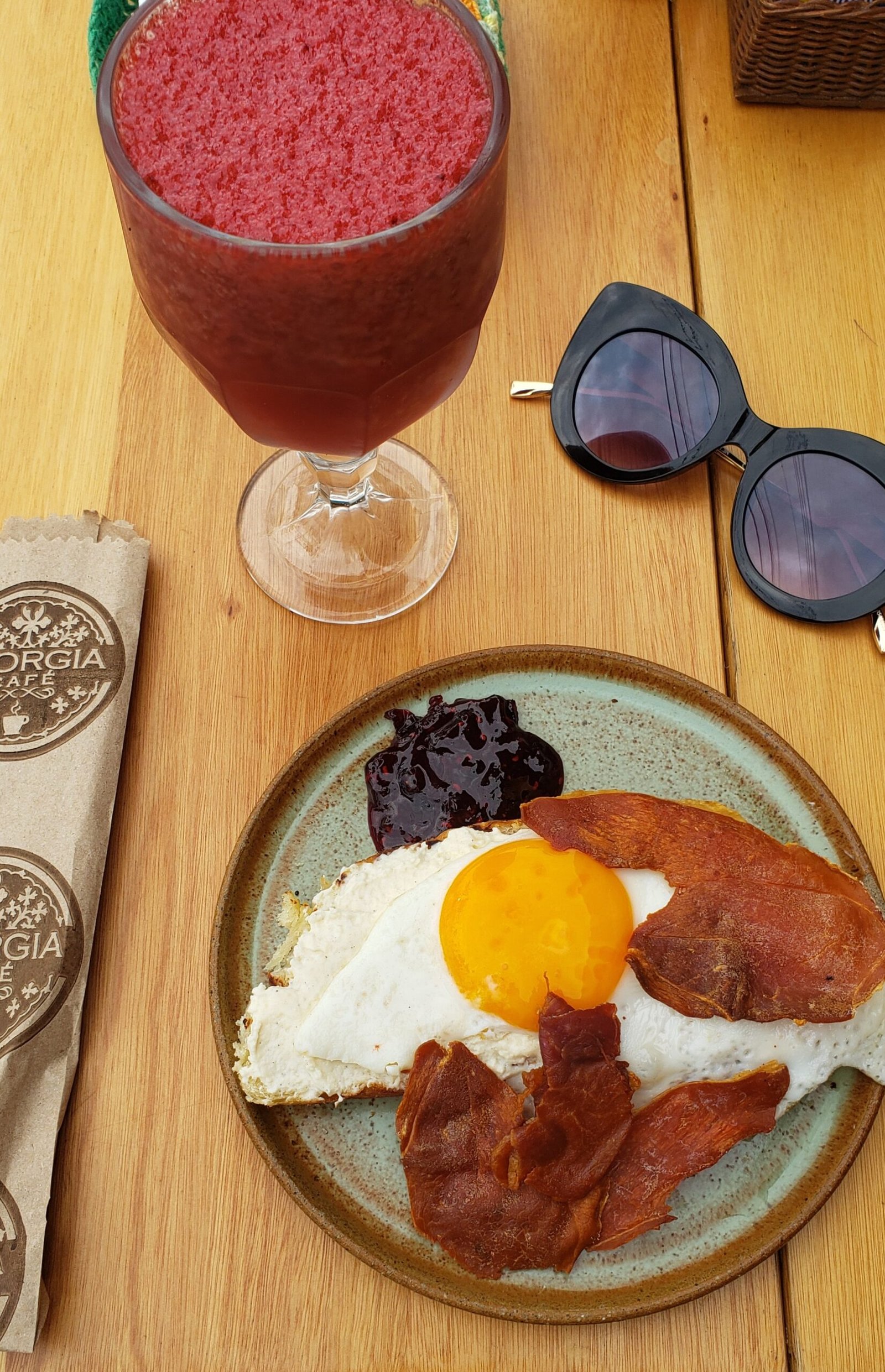 Mesa de madeira com prato verde de cerâmica com toat com ovo frito, bacon e geleia de amora, uma taça de suco de frutas vermelhas e um óculos escuro no canto superior direito.