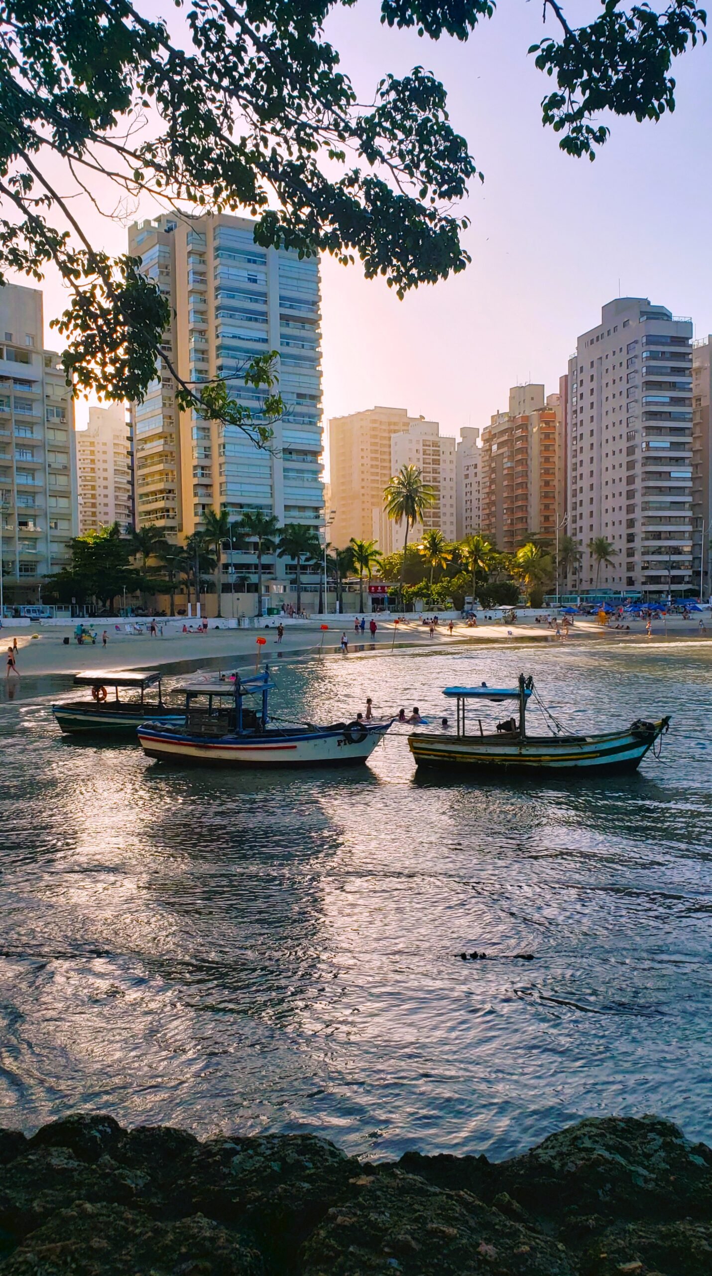 Fim da tarde na Praia das Astúrias, visto do canto dos pescadores, com o Sol se pondo entre os prédios da orla, o mar refletindo o céu e alguns barcos de pescadores no primeiro plano