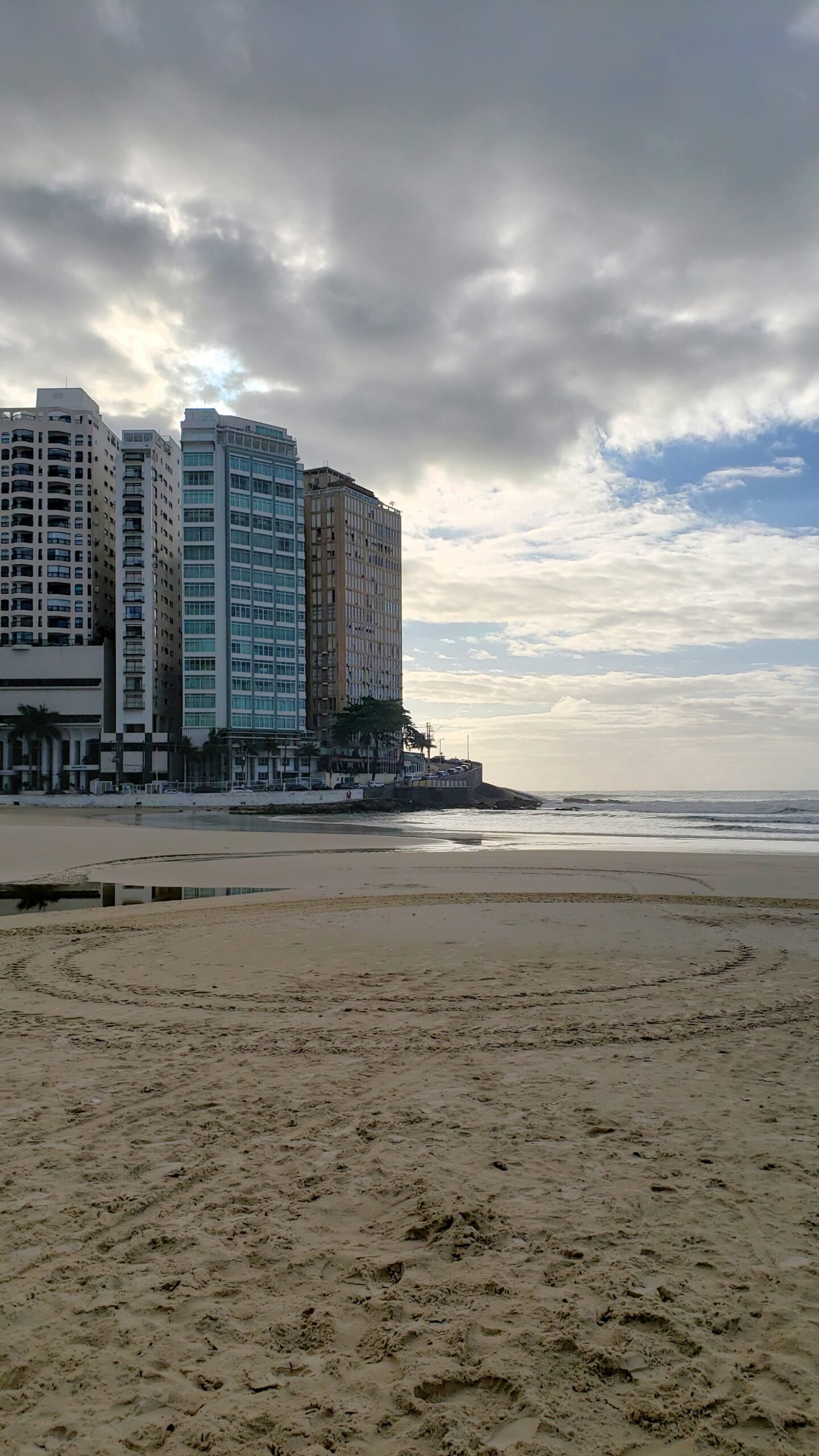 Canto da Praia de Pitangueiras no Guarujá, com prédios no canto direito, o mar no canto inferior direito e nuvens escuras no céu.