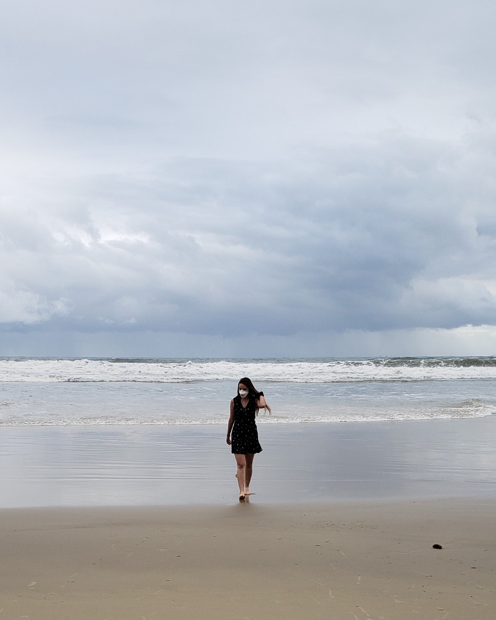 Mulher no centro, usando vestido e máscara, na praia na pandemia. Foto tirada na Praia de Pitangueiras no Guarujá